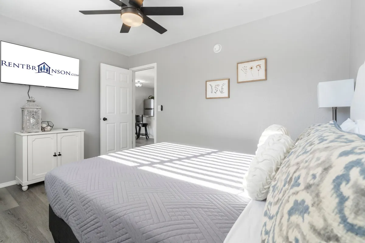 A queen-sized bed with a gray comforter is complemented by a ceiling fan, wall-mounted TV, tasteful wall art, and a nightstand, illustrating a cozy bedroom perfect for unwinding at the end of the day.