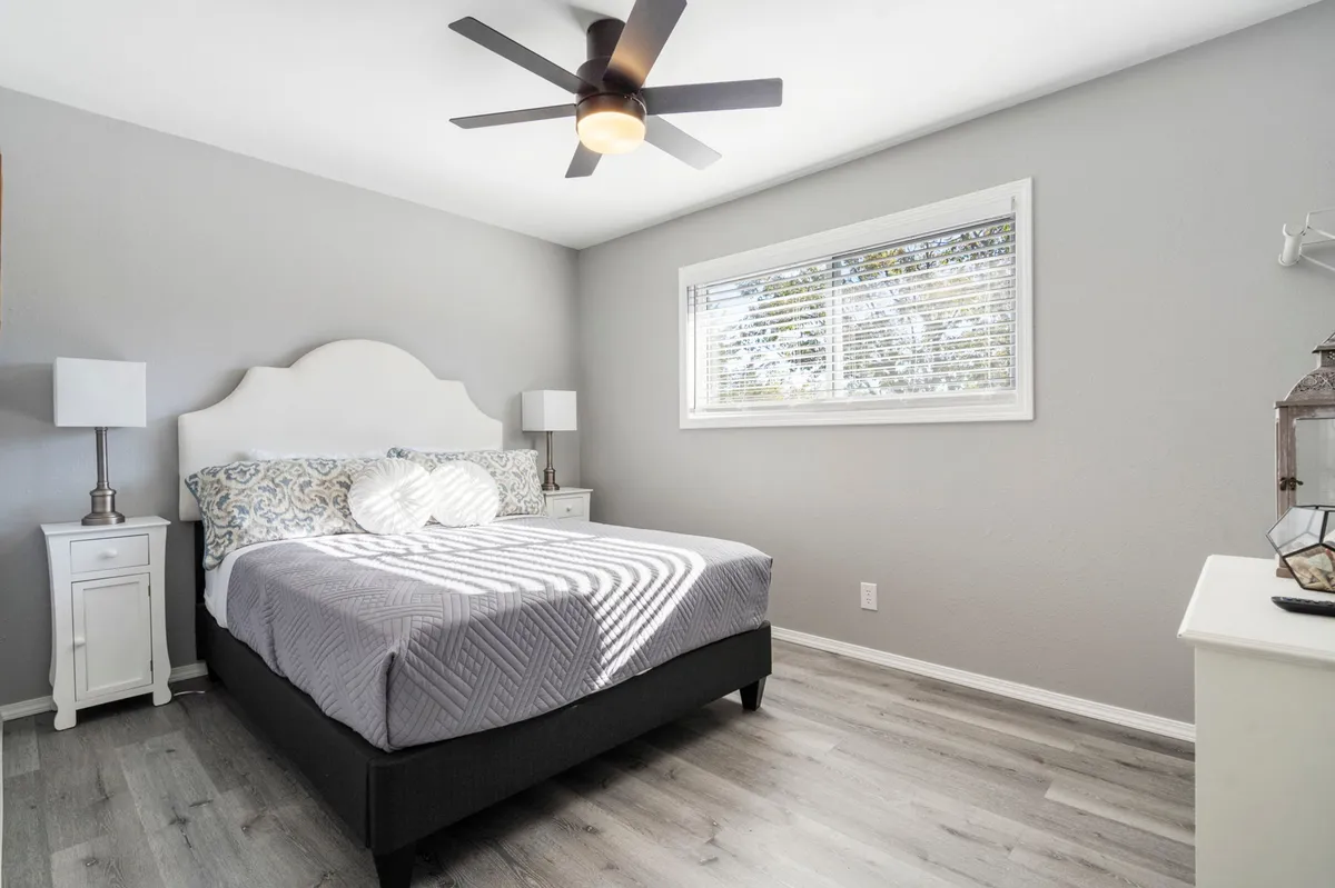 A queen bed adorned with decorative pillows sits in a softly lit bedroom. Natural light pours in through a window, illuminating side tables with lamps and inviting textures, creating a calming, inviting ambiance for relaxation or reading.
