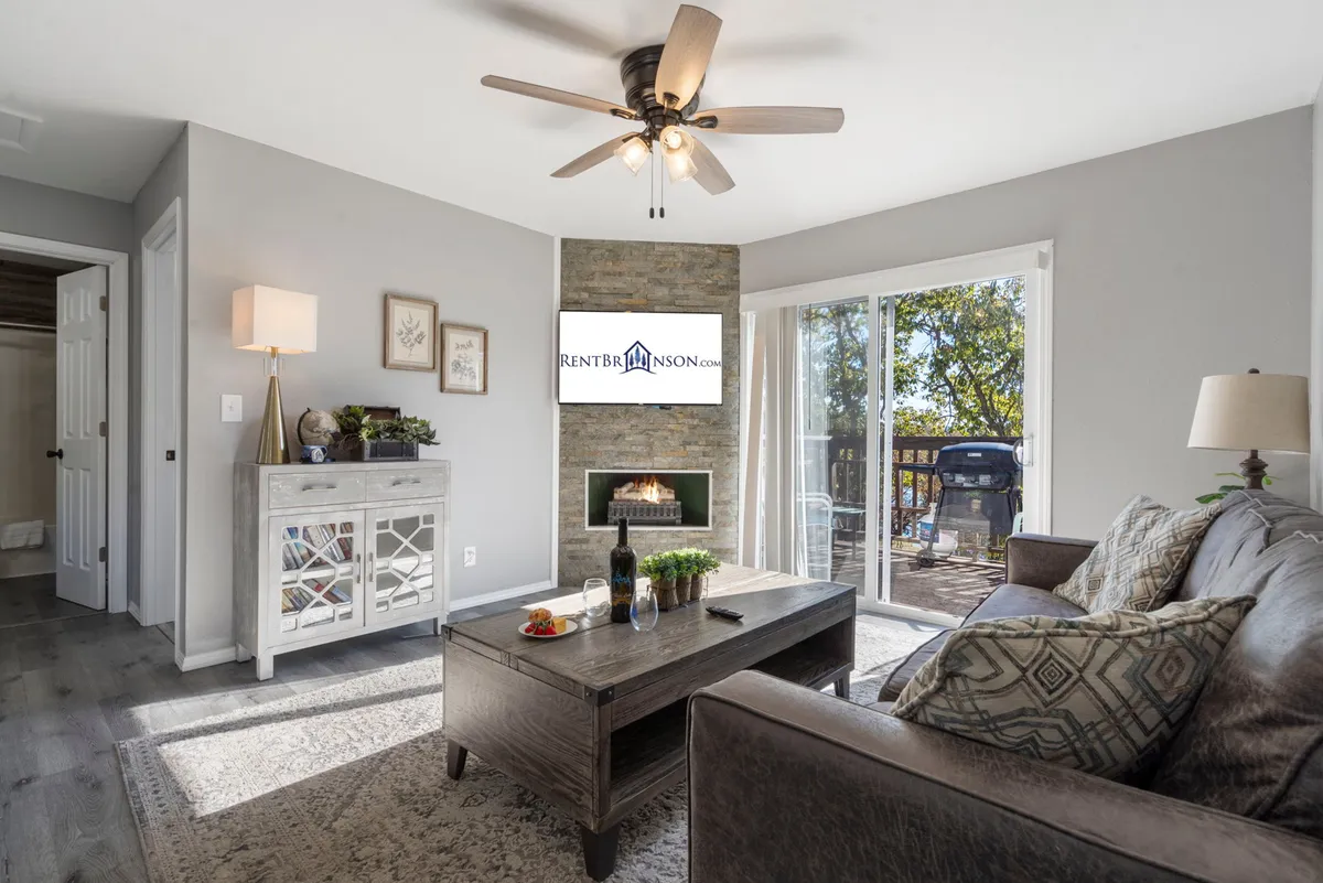 A comfortable living area featuring a plush sofa, a cozy fireplace, a streaming-ready TV, a coffee table, and a decorative lamp. Sunlight streams in through large windows, suggesting a bright, inviting atmosphere ideal for relaxing or entertaining.