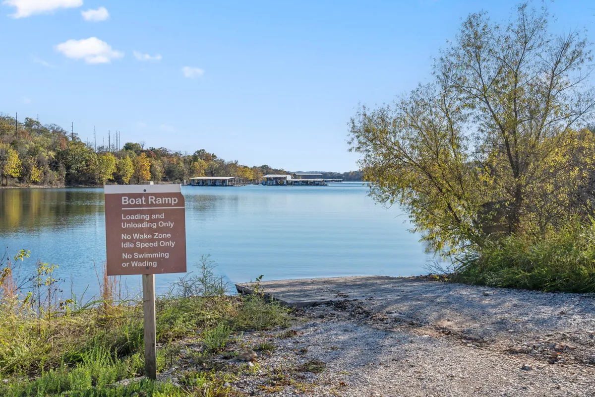 A picturesque view of a boat ramp gently sloping into calm lake waters, surrounded by trees and clear signage. The scene invites visitors to launch their boats or settle in for a peaceful fishing session, emphasizing easy lake access.