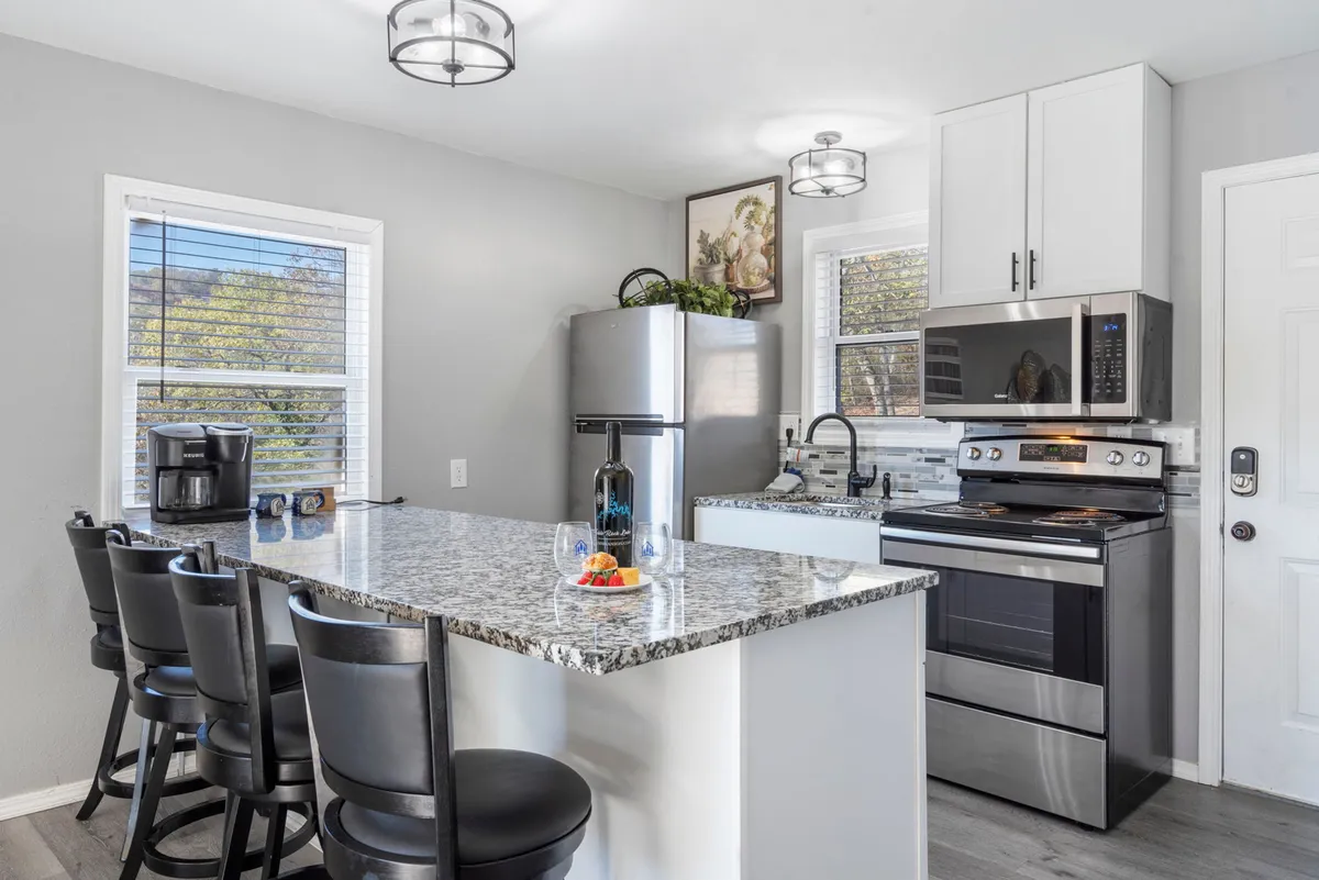The image showcases a stylish and inviting kitchen with gleaming granite countertops, modern appliances, and comfortable bar stools. A bottle of wine with glasses is artfully arranged, and framed artwork adds warmth, creating a welcoming space perfect for gathering, cooking, and lingering conversations.