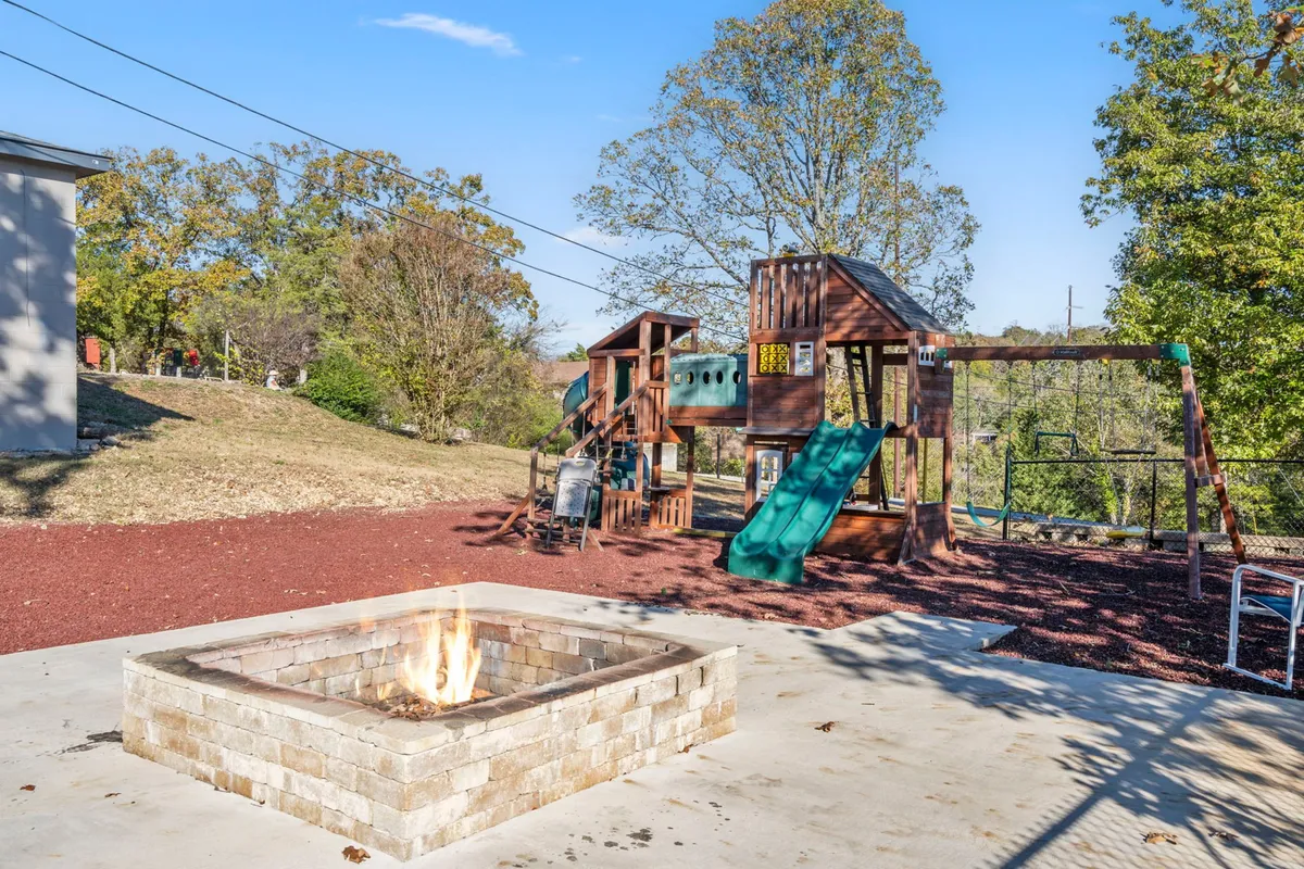 A vibrant outdoor area featuring a cozy fire pit surrounded by seating, with the on-site playground equipment visible in the background. Lush greenery frames the scene, inviting families and children to gather, play, and unwind together in a joyful and relaxed setting.