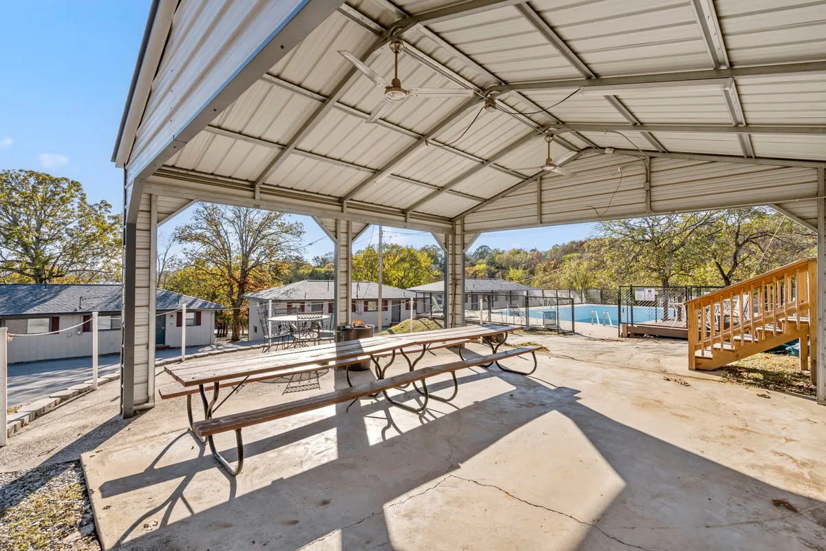 An open picnic pavilion with a large table under a wooden roof, surrounded by trees. The space is designed for group gatherings, shaded relaxation, outdoor meals, and socializing in a friendly, comfortable setting.